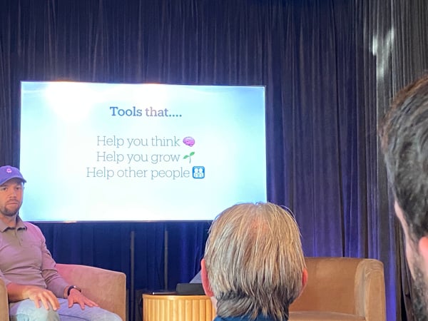 A photograph showing a presentation stage with a screen displaying text about tools that help people think, grow, and help others, with a speaker seated on the left and audience members in the foreground.