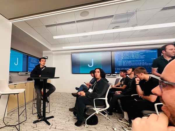 A candid photograph taken indoors showing a technical presentation in a modern office space, featuring a presenter sitting at a high table with a laptop addressing a small audience, with code and a large 'J' displayed on dual wall screens.