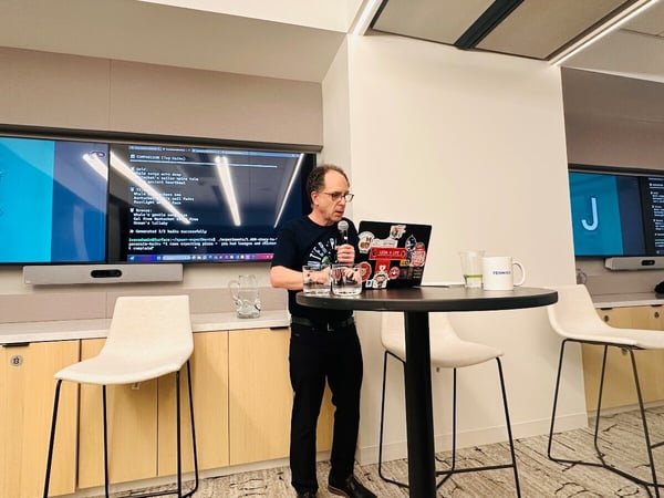 A man is giving a technical presentation in a modern office or conference room, standing behind a high table while speaking into a microphone and referencing his open laptop. Two large wall-mounted screens behind him display code output and a prominent letter 'J'.
