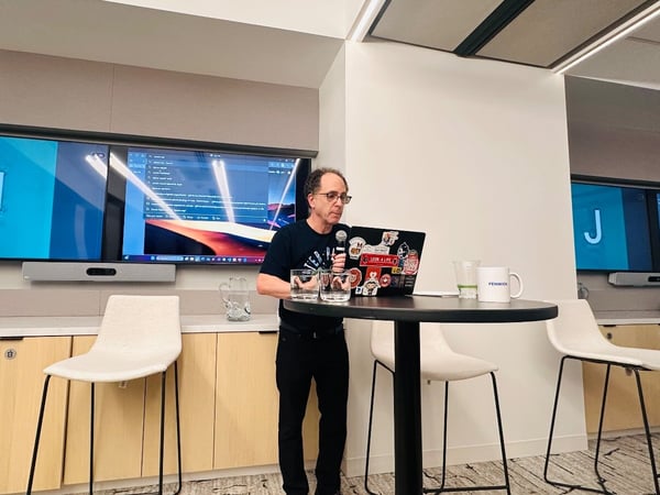 A man is giving a presentation in a modern office or conference room, standing behind a small round table with a laptop and a microphone. Behind him, two large wall-mounted monitors display a large letter 'J' on a blue background and some technical content.