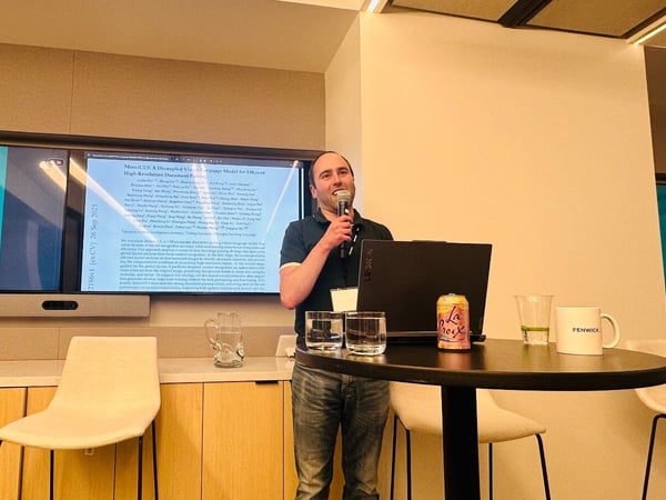 A man is giving a technical presentation indoors, holding a microphone, while standing next to a small table holding a laptop, drinks, and a mug branded 'FENWICK'. Behind him, a large screen displays a presentation slide containing the title and abstract of a document, alongside a large letter 'J'.
