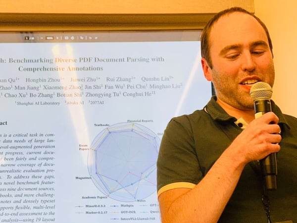 A man is giving a technical presentation, holding a microphone while speaking, with a large screen behind him displaying a research paper slide titled "OmniDocBench: Benchmarking Diverse PDF Document Parsing with Comprehensive Annotations."