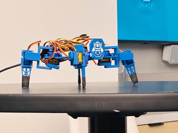 A close-up photograph of a small, blue, multi-legged robot prototype, featuring exposed wiring and servo motors, sitting on a dark table.