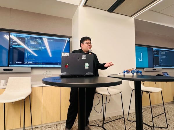 A man is giving a technical presentation or demonstration in a modern conference room, standing behind a high table with an open laptop and a small robotic device, while large screens behind him display code and the letter 'J'.