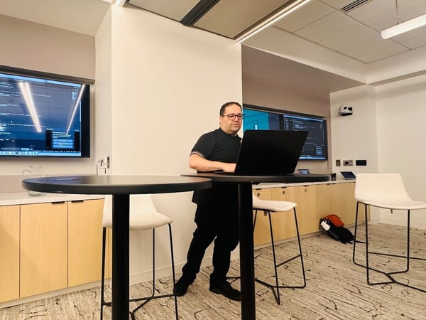 A photograph of a man standing at a high table, working on a black laptop in a modern conference room, with large wall-mounted screens displaying lines of code or configuration.