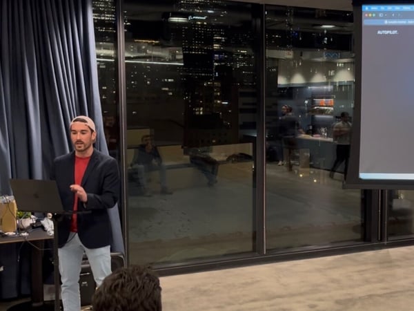 A photograph captures an indoor presentation event at night, showing a man presenting information displayed on a large screen against the backdrop of a city skyline visible through large windows.