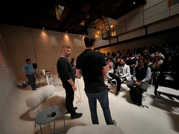 A low-light photograph captures an indoor event, showing two men standing in the foreground—one holding a microphone—addressing a large audience seated in rows. The event takes place in a spacious, industrial-style venue with exposed rafters and warm accent lighting on the back wall.