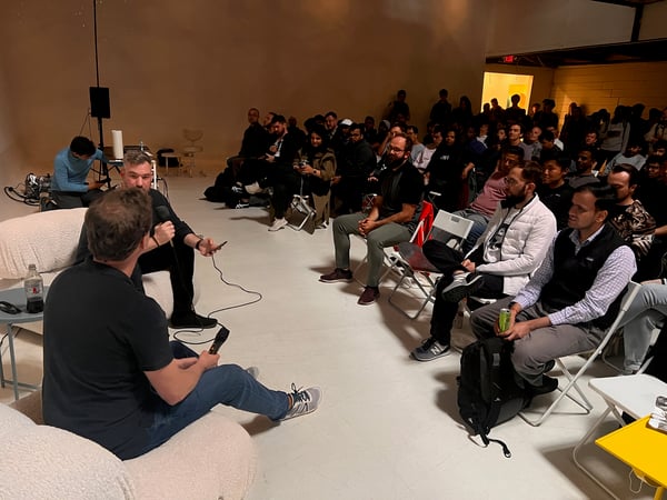 A photograph capturing an indoor event, likely a panel discussion or interview, where two men are seated and speaking into microphones while a large audience is gathered behind them. The setting appears to be a minimalist studio space with warm, focused lighting on the foreground subjects.