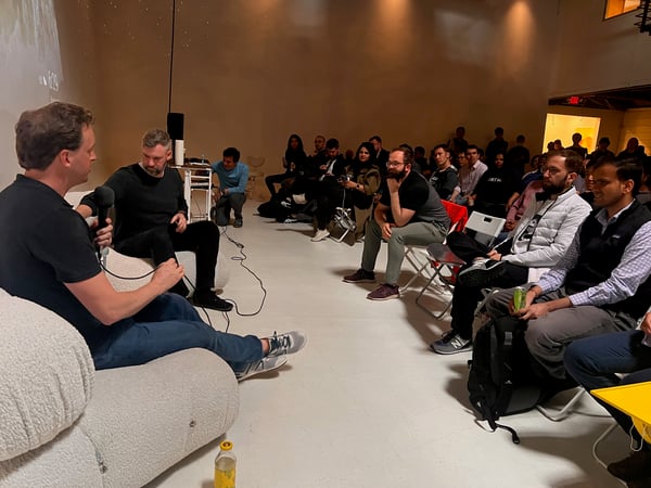 A photograph capturing an indoor professional event, likely a panel discussion or Q&A session, where two men are seated on a couch in the foreground, one holding a microphone, addressing a large audience seated on folding chairs.