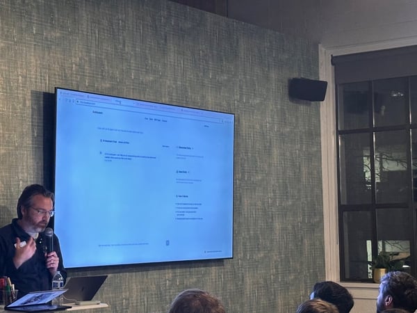 A man wearing glasses is giving a presentation at an indoor event, standing beside a large screen displaying a website or application interface, with an audience visible in the foreground.