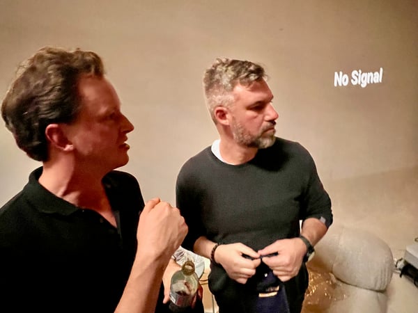 A low-light photograph capturing two men standing indoors, seemingly engaged in conversation, with a wall or projection screen behind them displaying the message "No Signal."