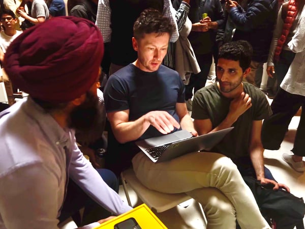 A candid photograph captures three men engaged in a discussion around an open laptop in a dimly lit, crowded indoor setting, likely during a networking or professional event.