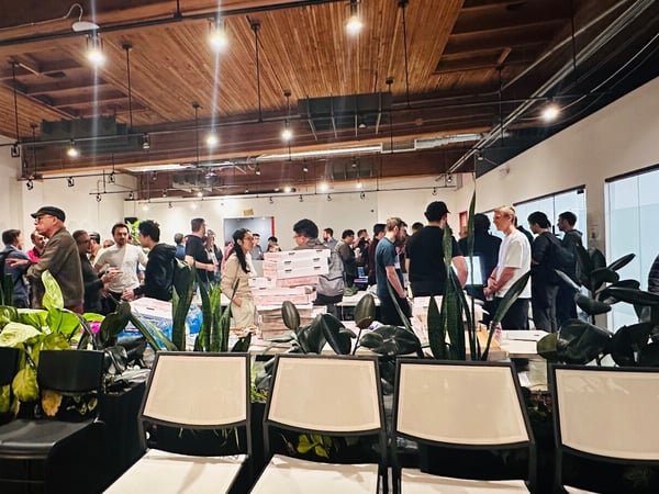 A candid photograph captures a large indoor gathering, likely a corporate or tech meetup, where a crowd of people is mingling beneath a wooden-beamed ceiling and bright track lighting.