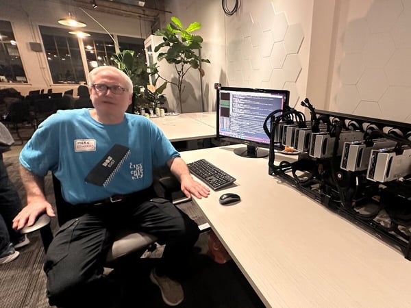 A man wearing a blue t-shirt and glasses is seated at a desk next to a large, open-frame computer rig (likely for cryptocurrency mining or HPC) and a monitor displaying code in a modern office environment.