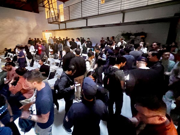 A candid photograph captures a large crowd of people socializing and mingling in a dimly lit, high-ceilinged indoor venue, characterized by industrial architecture and rows of white folding chairs.
