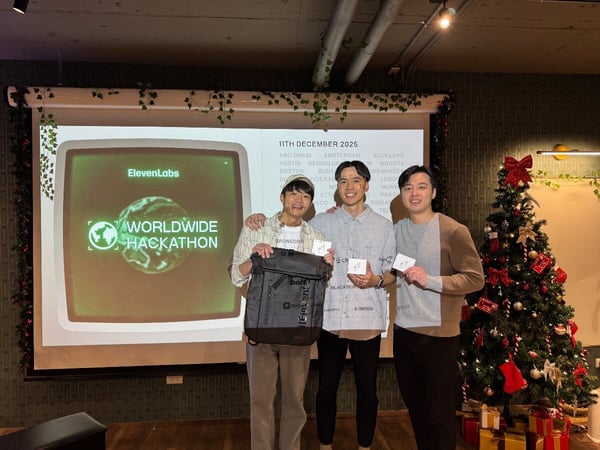 Three young men pose for a photo indoors, holding small white cards and a backpack prize, standing in front of a projection screen advertising the ElevenLabs Worldwide Hackathon and a decorated Christmas tree.
