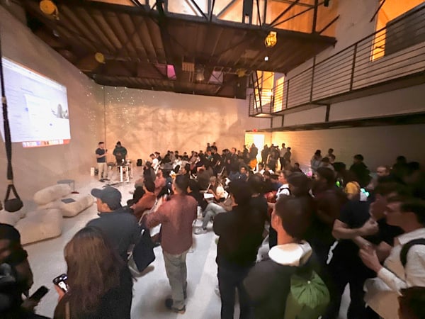 A photograph captures a large indoor gathering, likely a presentation or tech event, held in a spacious venue with industrial architecture.