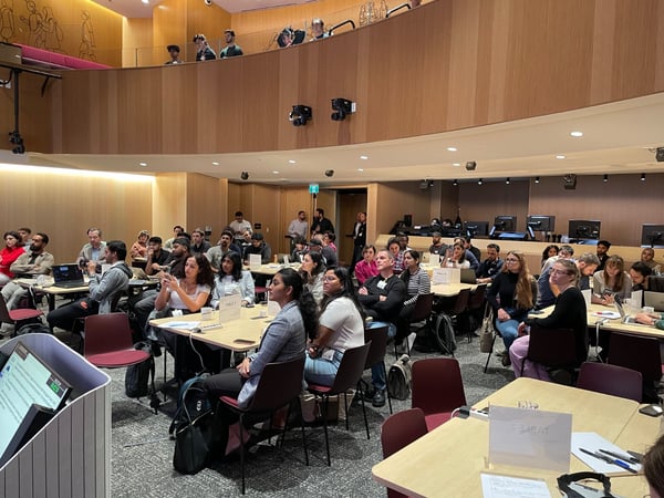 A photograph capturing a large audience seated at tables in a modern, wood-paneled lecture hall or conference room, attending a presentation or workshop.