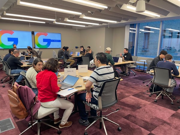 A photograph capturing a group of approximately 16 people seated at modular tables in a modern conference room, actively working on open laptops during what appears to be a workshop or meeting.