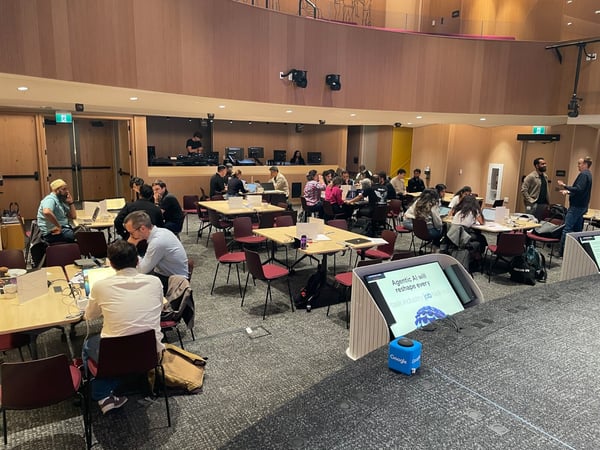 A candid photograph capturing a large group of people attending a workshop or hackathon, collaborating at square tables with laptops in a modern auditorium setting with wood-paneled walls.