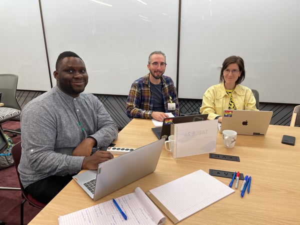 Three colleagues, two men and one woman, are seated at a light wooden conference table with laptops and notebooks, smiling directly at the camera in a modern office meeting room.