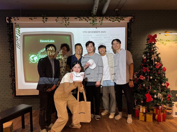 A group of seven people, primarily young adults, pose for a photo in front of a projection screen displaying "ElevenLabs" content and a fully decorated Christmas tree, likely at a holiday event. The woman in the foreground holds up branded merchandise while the group stands in a festive indoor setting.
