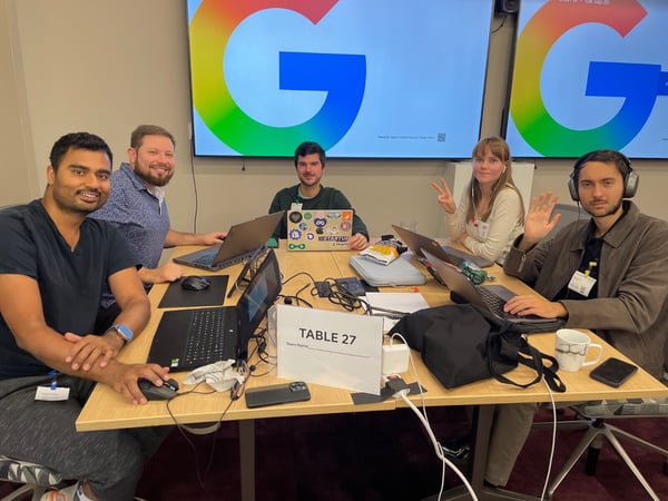 A group of five participants, four men and one woman, are gathered around a table with laptops, working collaboratively during an event, featuring large screens displaying the colorful Google 'G' logo behind them.