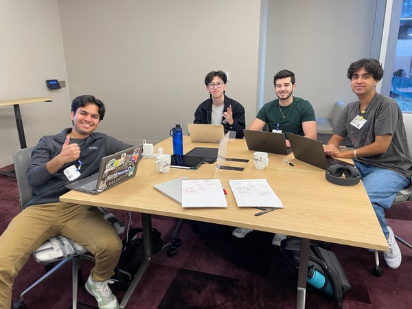 Four young men are seated around a light wooden table in a modern office setting, working on laptops and collaborating during a group session.