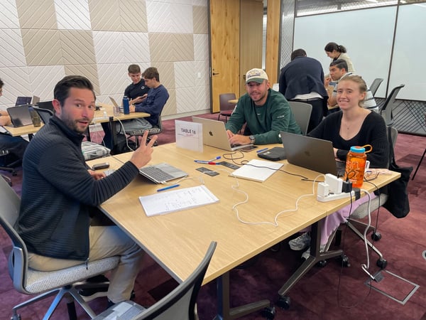 A candid photograph captures a group of people working collaboratively on laptops around large wooden tables in a modern office or study space, with the man in the immediate foreground smiling and giving a peace sign.