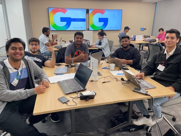 A candid photograph captures a group of young people working collaboratively on laptops around large tables in a modern office setting, with two large screens displaying the Google logo in the background.