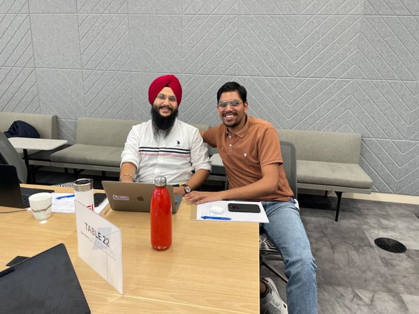 A photograph showing two men smiling while seated at a light wooden table in a modern office or co-working space, surrounded by laptops and work materials.