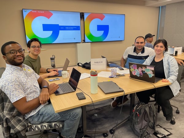 A group of four people are seated around a conference table working on laptops in a brightly lit indoor setting, with two large screens behind them displaying the colorful Google logo and search interface.