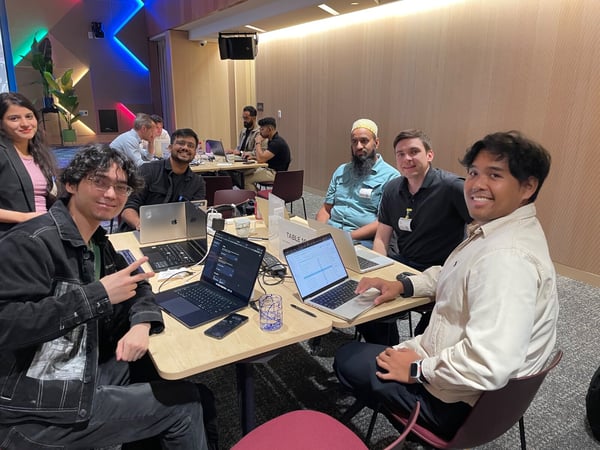 A group of diverse individuals smiles while sitting around a table with open laptops in a modern event space, suggesting participation in a collaborative workshop or hackathon.