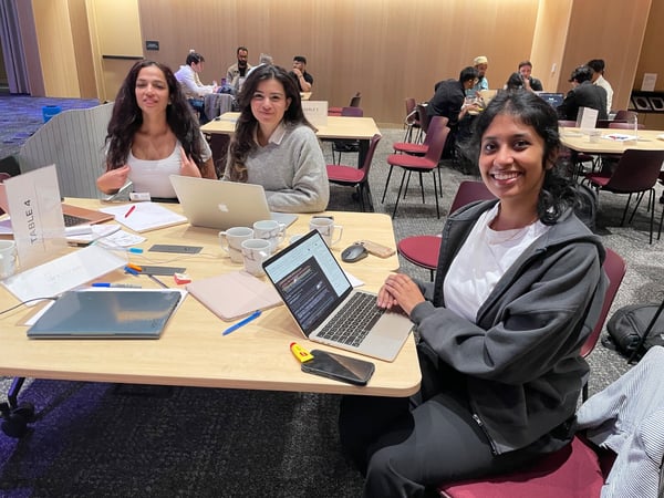 Three young women are seated at a wooden table with laptops and papers, smiling directly at the camera in a brightly lit, modern conference or workshop setting.