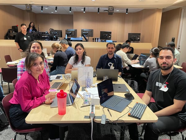 A group photo taken indoors during a workshop or coding session, showing five individuals seated at a light wooden table looking toward the camera, surrounded by open laptops and accessories. Several other participants are visible working at tables in the background, set against wood-paneled walls and multiple computer monitors.