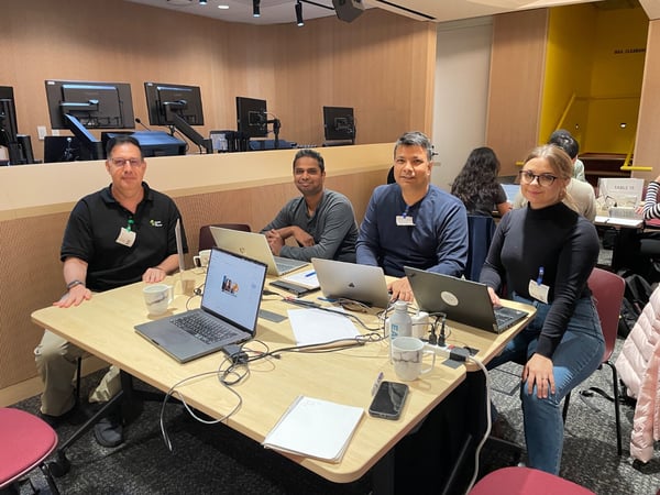A group of professionals is seated around a table in a modern conference or training room, working on laptops during what appears to be a workshop or meeting. Four individuals are prominently featured, looking toward the camera.