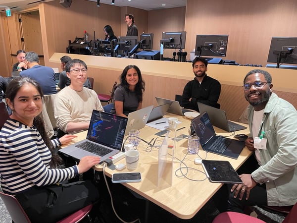 A group photo of five young professionals—three men and two women—smiling directly at the camera while sitting around a table with open laptops, likely at a coding workshop or conference.