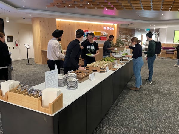 A candid photograph showing a group of people serving themselves food from a buffet line set up in a modern indoor office or event space, featuring light wood paneling and a sign reading "hi there" in the background.