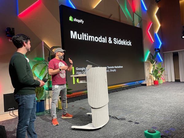 A photograph capturing an indoor presentation where two men are on stage in front of a large screen displaying the title 'Multimodal & Sidekick' and the Shopify logo. The stage features a modern podium and a geometrically patterned wall illuminated by colorful neon lights.
