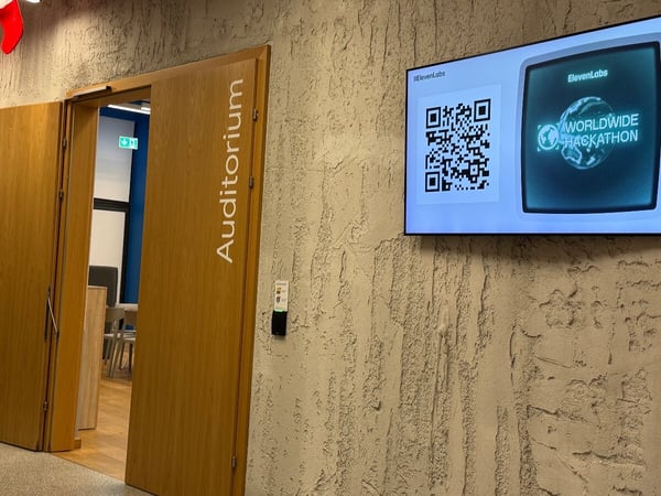 A photograph showing the entrance to an Auditorium, featuring a wooden door labeled vertically, next to a textured beige wall where a large digital screen displays event information for an ElevenLabs WORLDWIDE HACKATHON and a corresponding QR code.