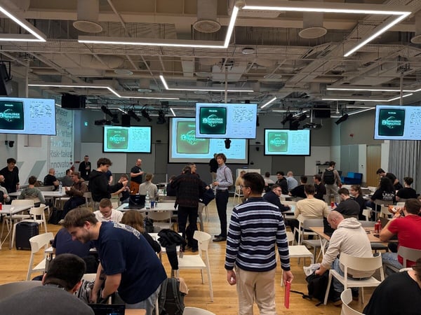 A wide-angle photograph capturing a busy indoor event, likely a hackathon or workshop, where numerous participants are seated at white tables using laptops and engaging in discussions in a large, modern hall.
