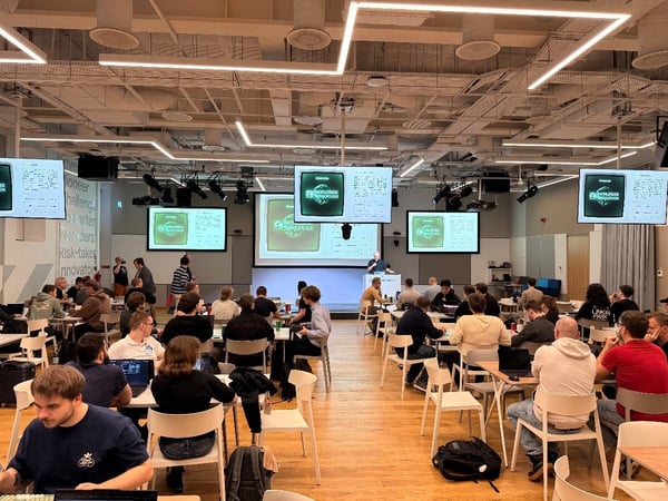 A wide-angle photograph captures a large group of people seated at tables in a modern, brightly lit indoor event space, participating in what appears to be a workshop or hackathon. Multiple screens display presentation slides to the audience.