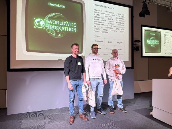 A photograph taken at an indoor event, showing three men standing side-by-side in front of a large projection screen that displays details for the 'ElevenLabs WORLDWIDE HACKATHON,' including the date, participating cities, and sponsors.