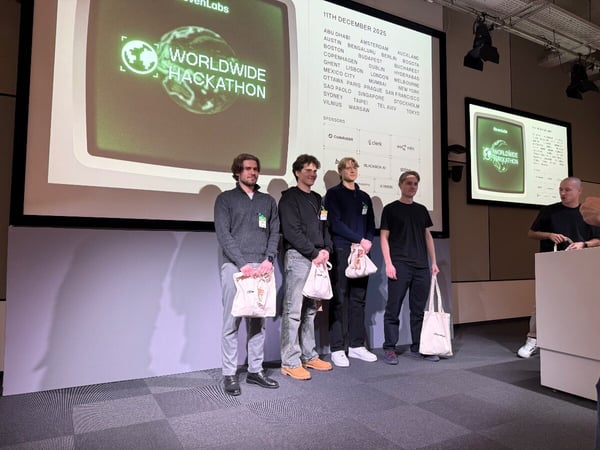 A photograph captures four young men standing on a stage during an event, presumably winners or participants of a hackathon, holding white tote bags, with large screens behind them displaying event details and sponsors.