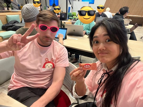 A playful photo of a man and a woman in pink shirts posing for a selfie indoors, with the woman holding a KitKat bar, while several other people work on laptops in the background.