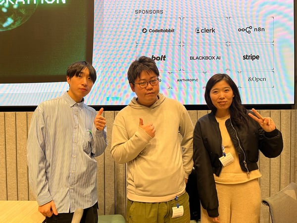 A group photo featuring three individuals (two men and one woman) posing indoors in front of a large screen displaying a list of sponsors for an event.