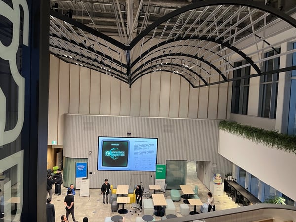 A high-angle photograph captures a modern, multi-story corporate atrium or event space featuring vaulted black metal arches overhead and light-colored walls, where a hackathon event is taking place. A large screen displays the title 'WORLDWIDE HACKATHON' hosted by ElevenLabs, and several people are standing and mingling among modern seating arrangements below.