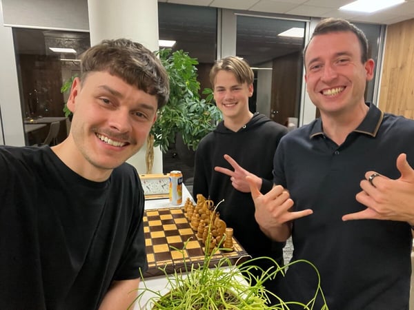 A casual photo showing three smiling men posing together indoors, with a chessboard, chess clock, and potted plants visible in the foreground.
