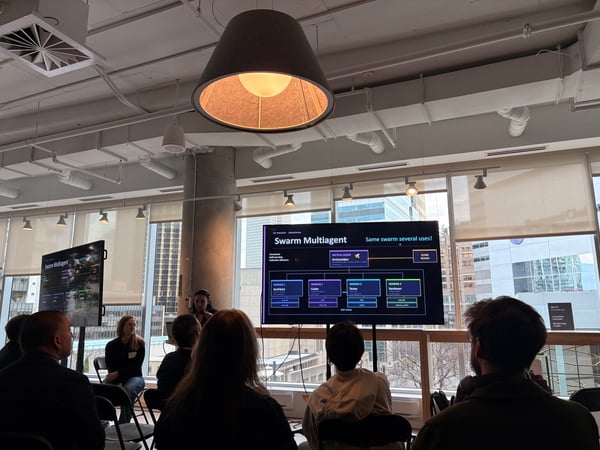 A group of people are attending a tech presentation in a modern office space with large windows. The main screen displays a diagram about 'Swarm Multiagent' systems.
