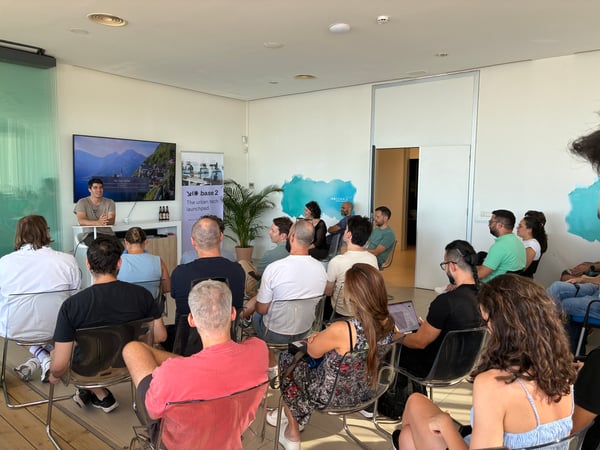 A group of people are seated in a modern indoor room, attending a presentation delivered by a speaker standing near a television screen.
